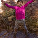 A girl standing in a muddy stream with her arms outstretched.