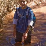 A boy standing in muddy water with a backpack.