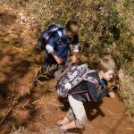 Two boys running through a muddy trail with backpacks.